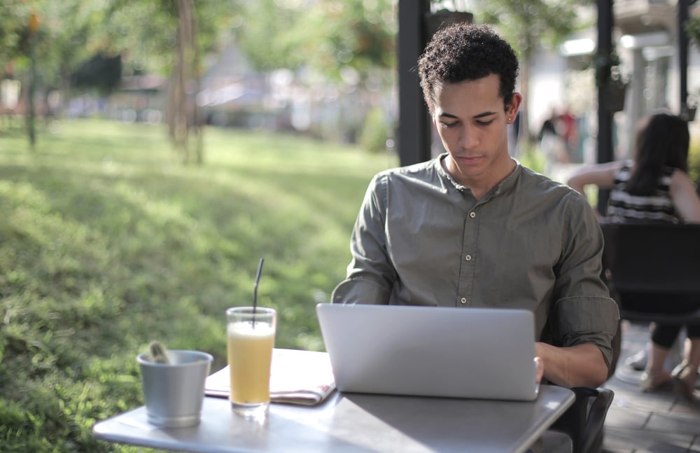 A male-presenting person works on a laptop at an outdoor cafe.