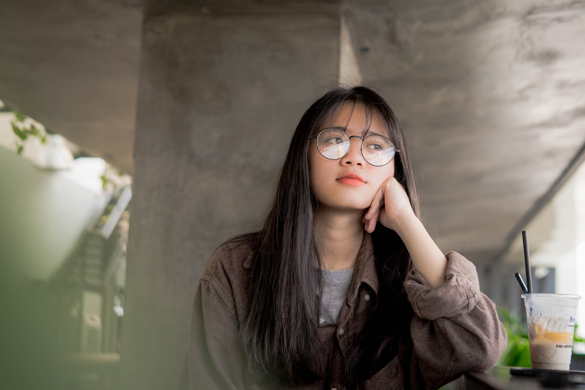 A female-presenting person wearing glasses sits in a cafe and looks off into the distance, waiting.