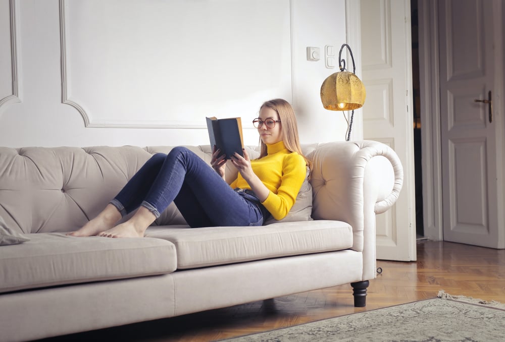 A female-presenting person in a bright yellow turtleneck and blue jeans sits on a couch reading the opening line of a book.