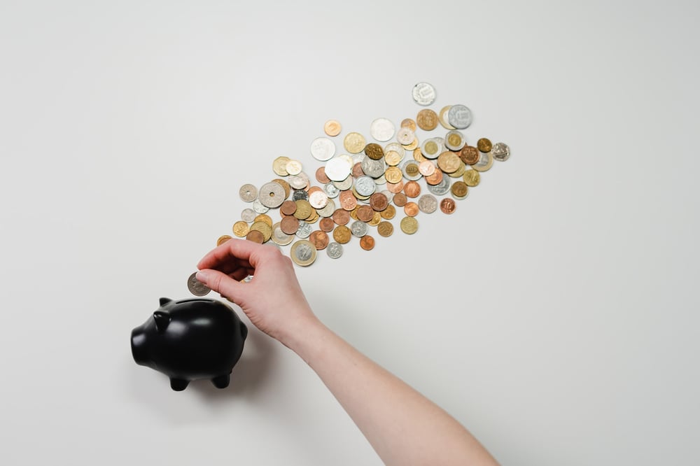 Coins scattered on a white surface. A hand puts one into a black piggy bank.