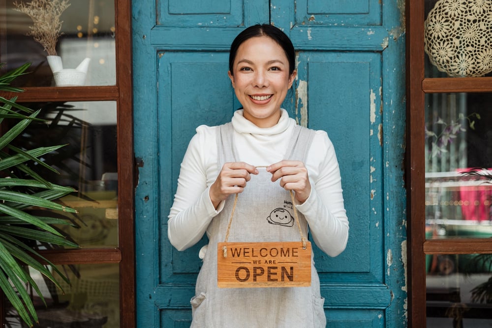 A female-presenting person stands in front of a shop smiling and holding an "open" sign.