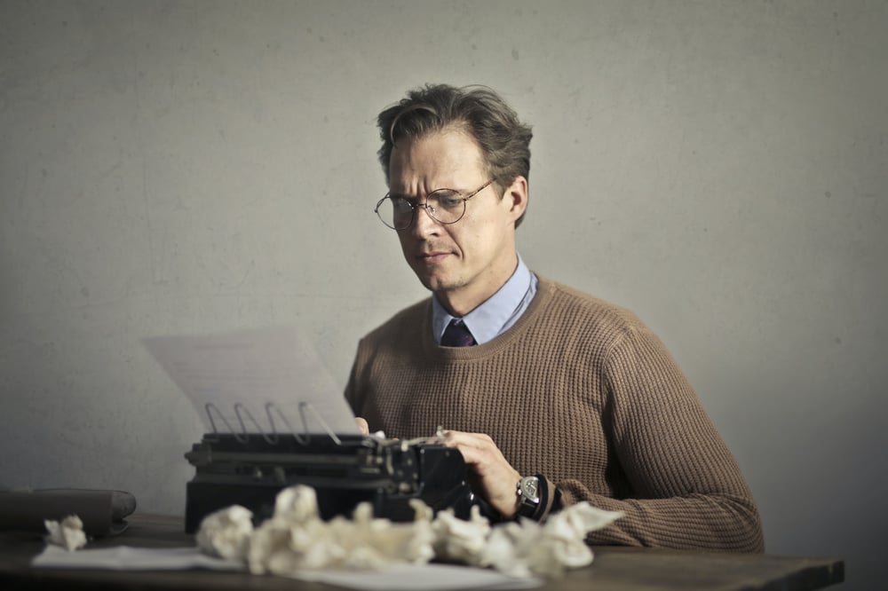 A male-presenting person types on a typewriter with a bunch of balled-up papers surrounding them.