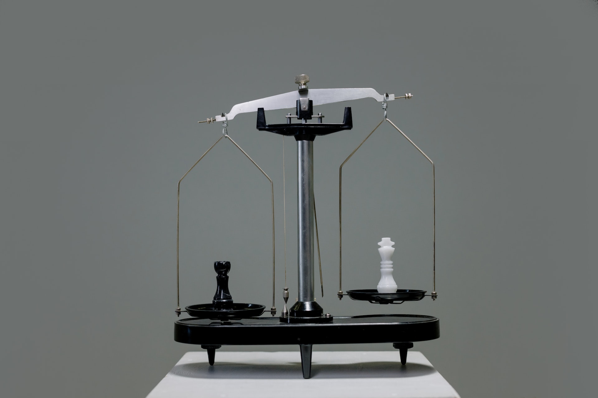 A scale with a black chess piece on one tray and a white one on the other.