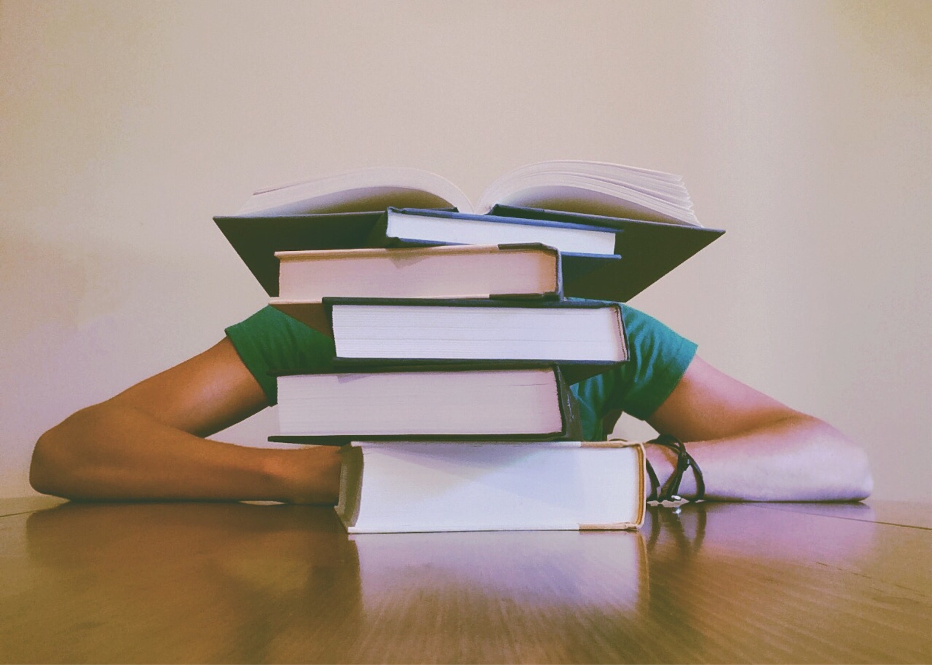 A person rests their head on the table, hidden behind a stack of research books.
