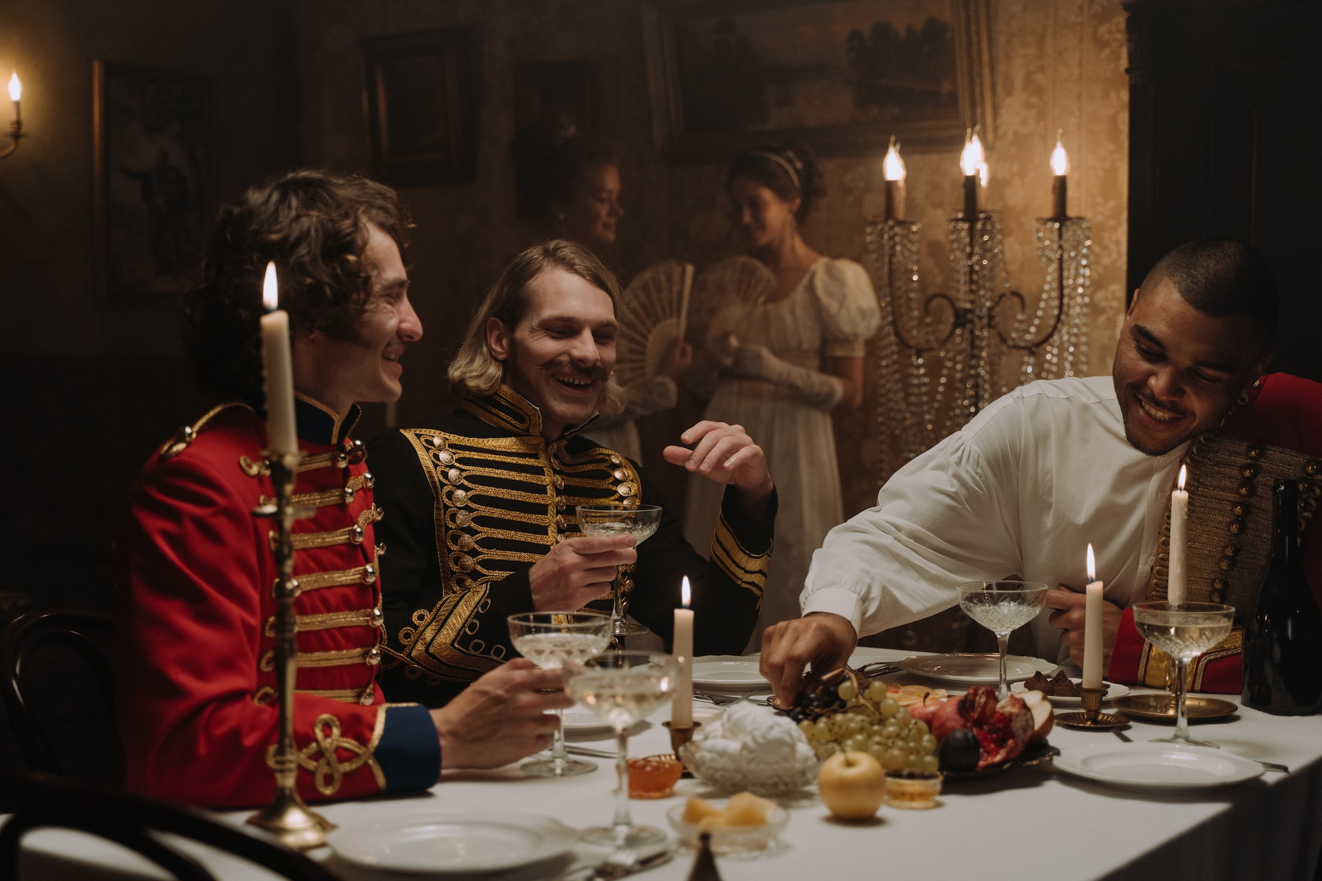 People dressed in 18th century British clothing laugh around a candlelit dinner table.