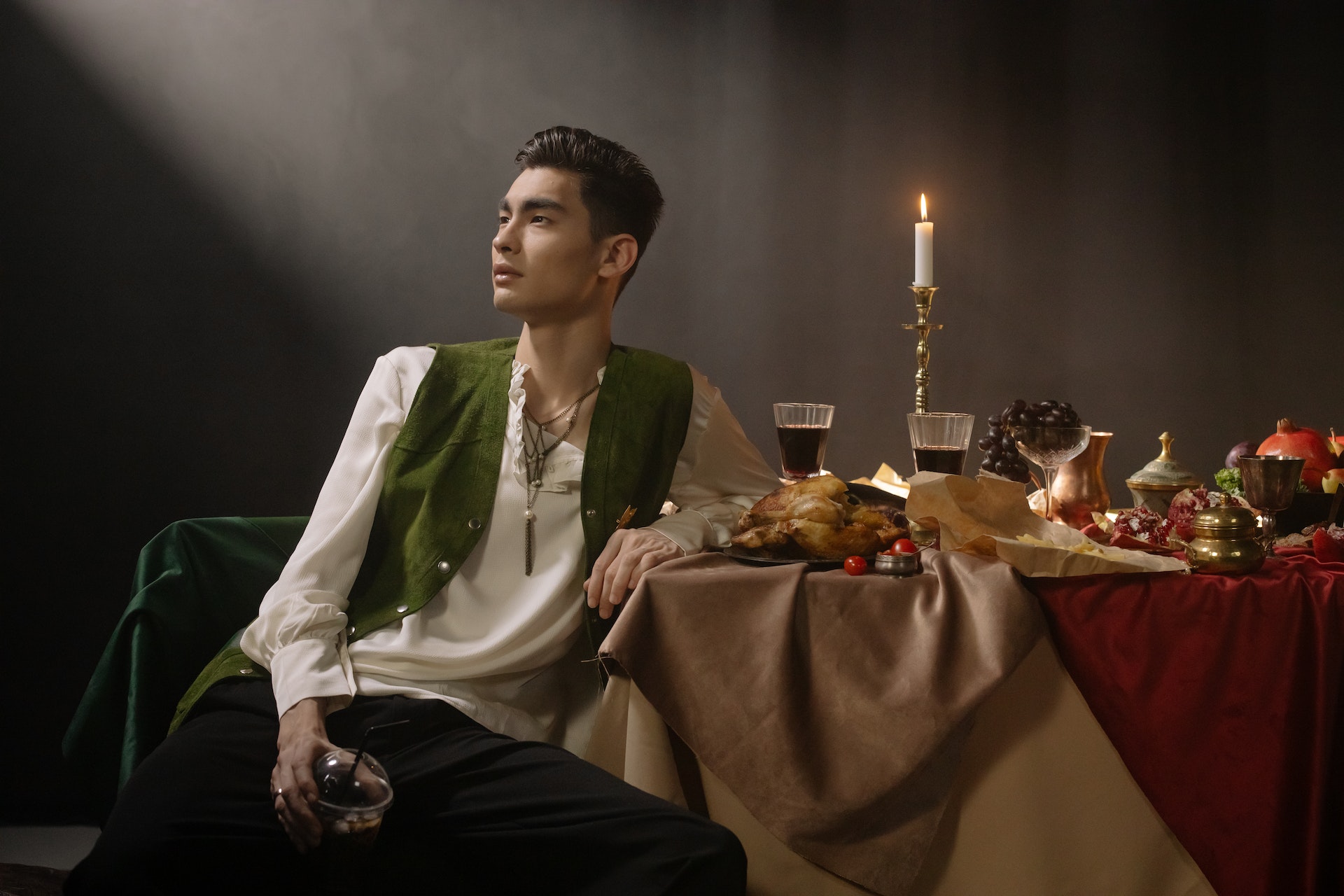 A person in Renaissance clothing sits at a table full of food and stares off into the distance.