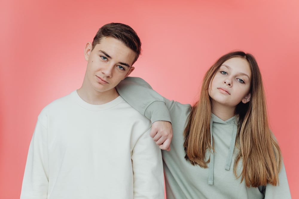 Two unsmiling teenagers pose together in front of a pink background.