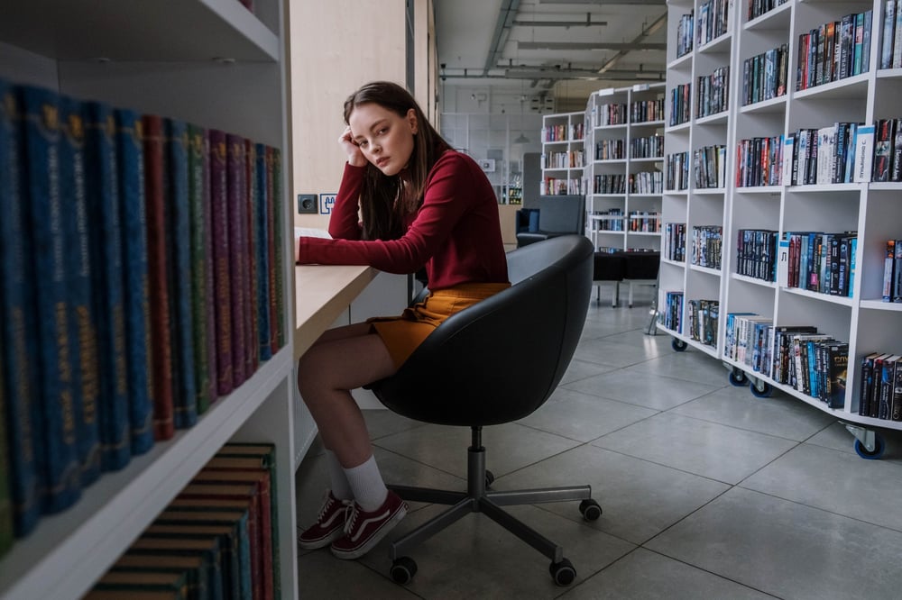 A stressed-looking teenager sits at a table in a library.