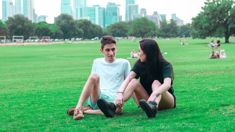 Two teenagers have a conversation while sitting on the grass in a city park.