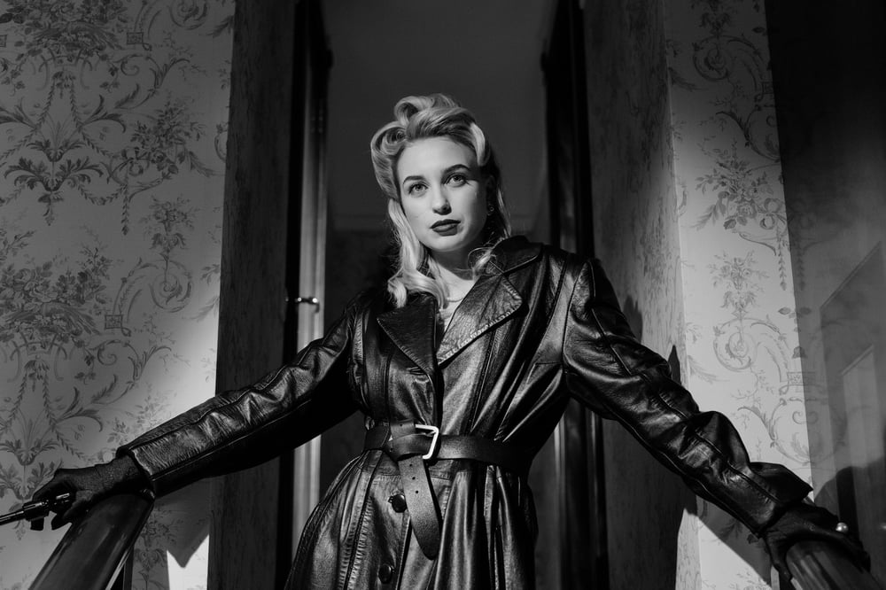 A female noir character in a black leather jacket stands in a doorway holding a gun.