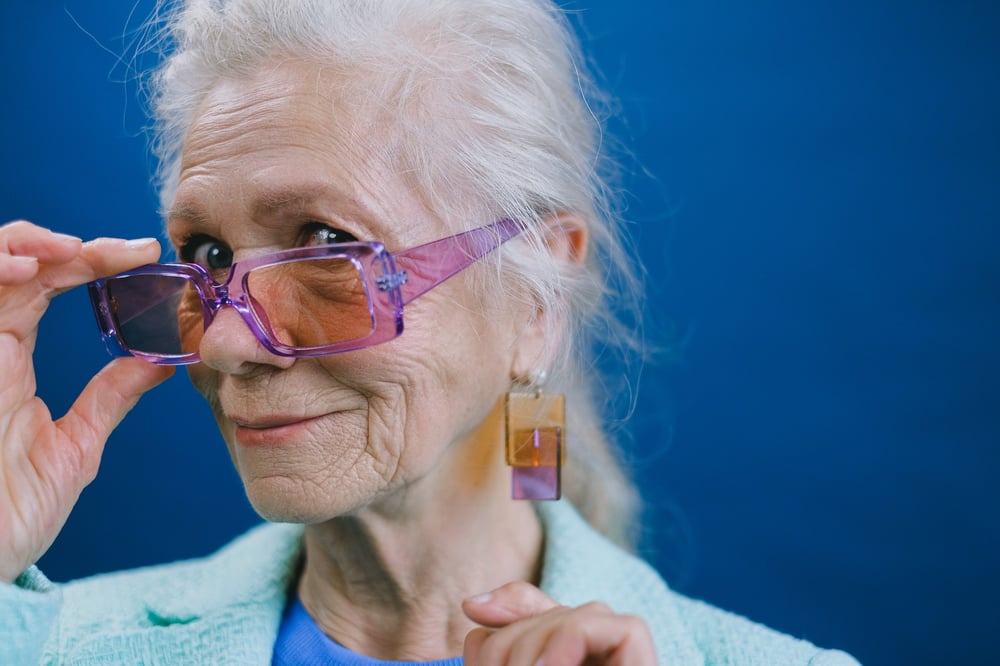 An older, female-presenting person looks over the top of their purple glasses.