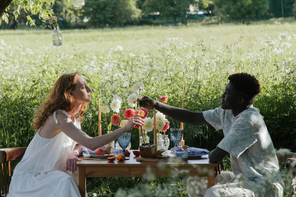 A couple eats together at a little table in a field of wildflowers.