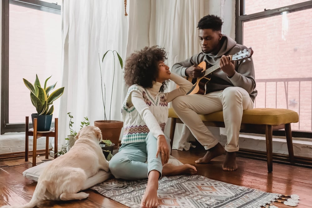 A couple sits in a living room playing guitar beside a dog.