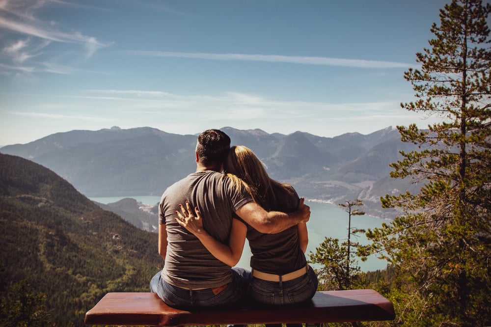 A couple sits on a bench in the mountains overlooking an alpine lake.