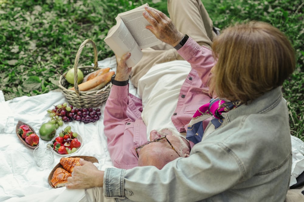 An older couple sits together on a picnic blanket reading a book.