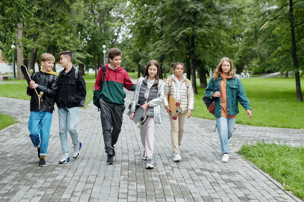 Six middle grade kids with backpacks and skateboards walk on a path through a park.