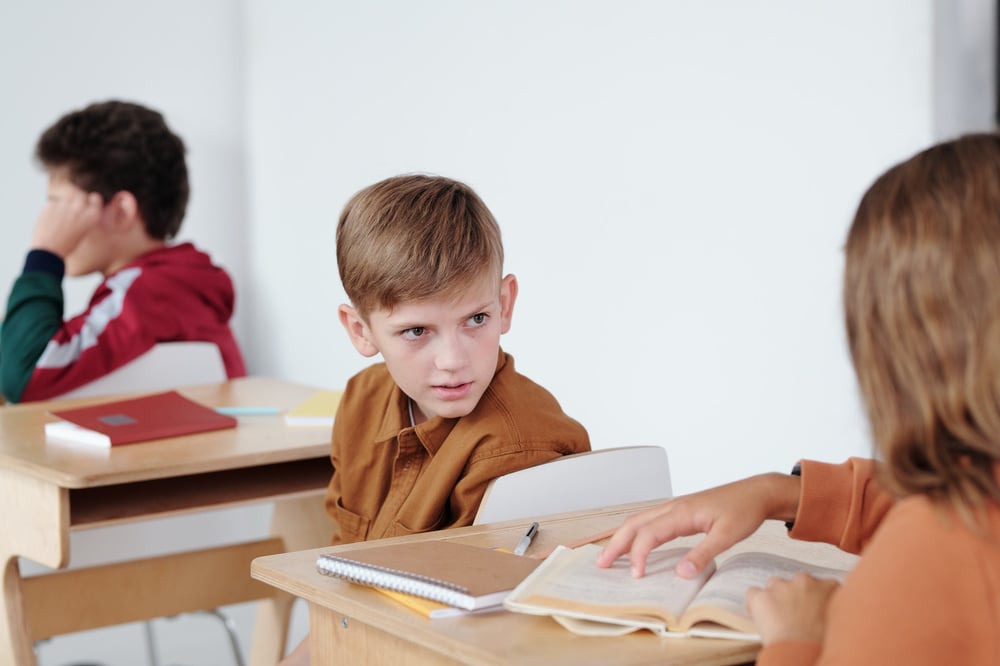 A middle grade kid at a school desk turning around to talk to the kid behind them.