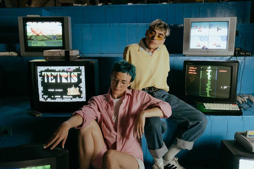 Two teenagers sit in a room in front of vintage computer screens with 1980s video games on them.