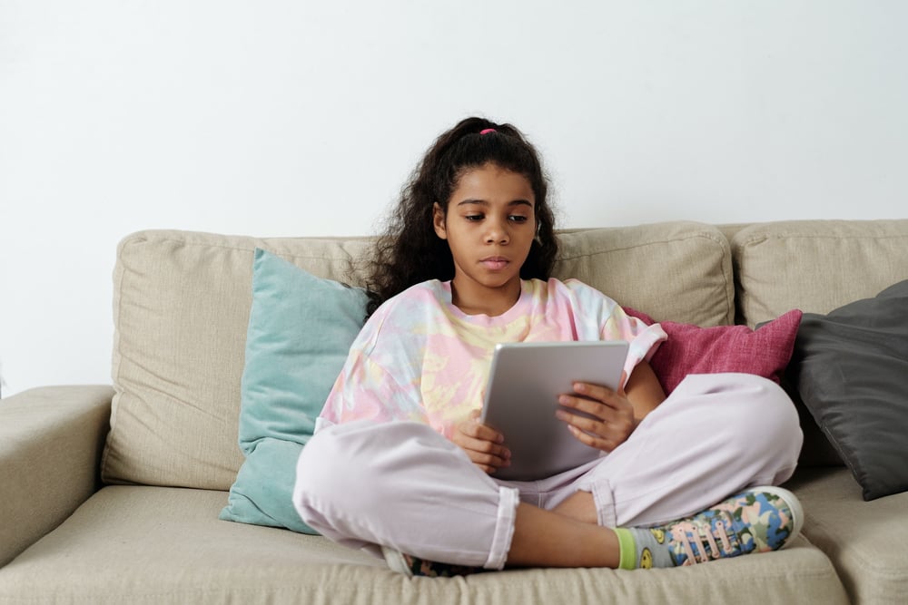 A teenager sits on a couch reading a novel on a tablet.