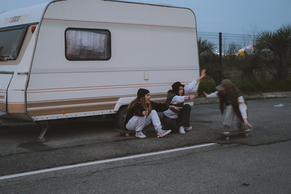 Teenagers skateboard beside a vintage camper.