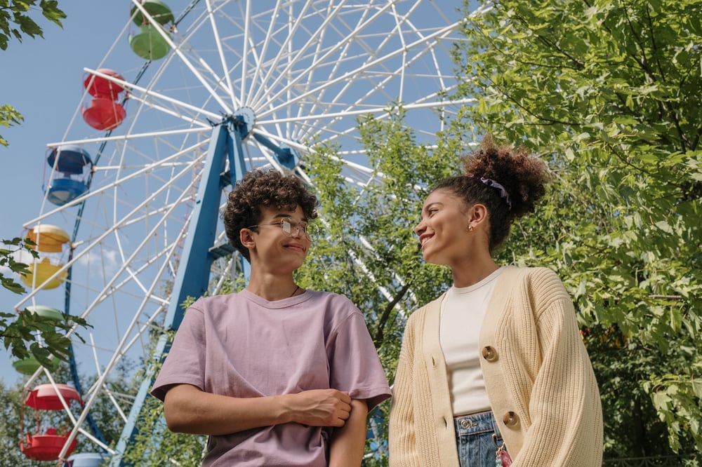 Two teenagers talk under a ferris wheel.