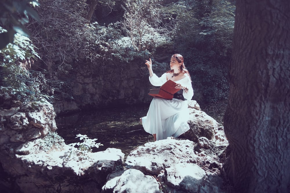 A teenager in a long, white dress sits beside a pond in the forest.