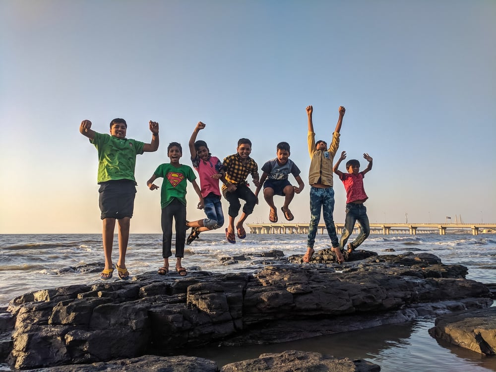 Seven middle graders jump on a rock in the ocean.
