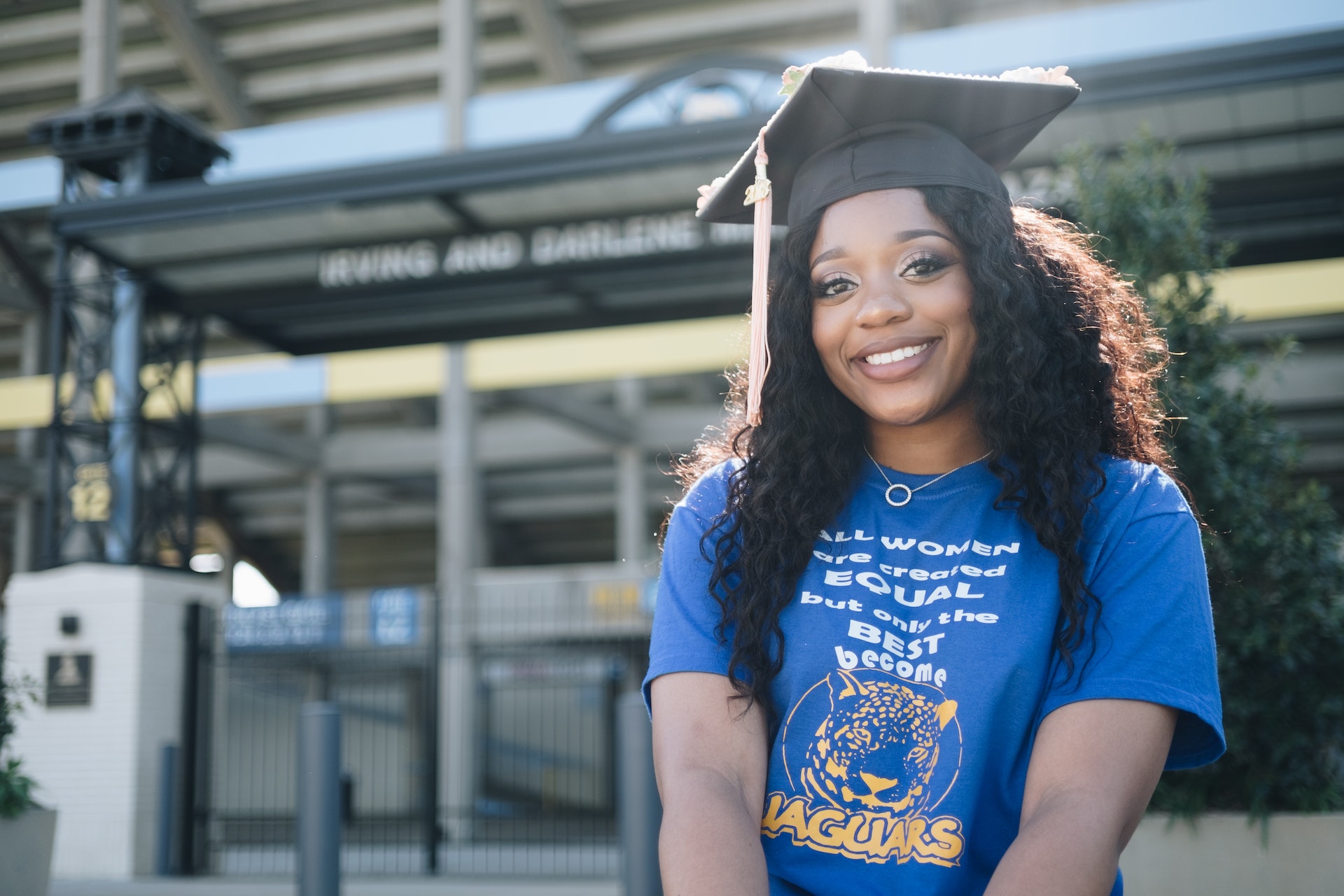 A person smiles in front of a building, wearing a graduation cap.