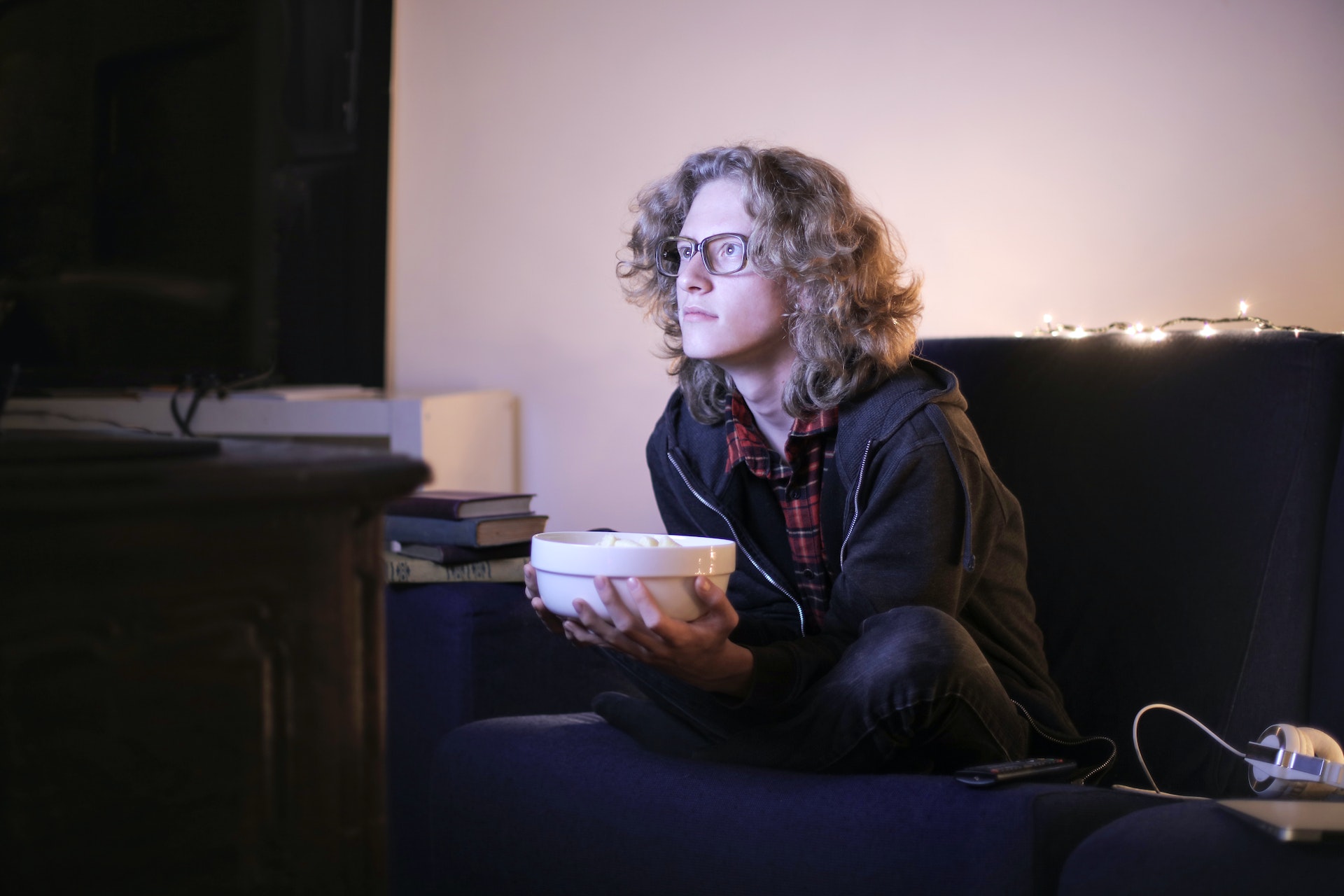 A person in glasses sits on a couch holding a big bowl and staring intently at a television screen.