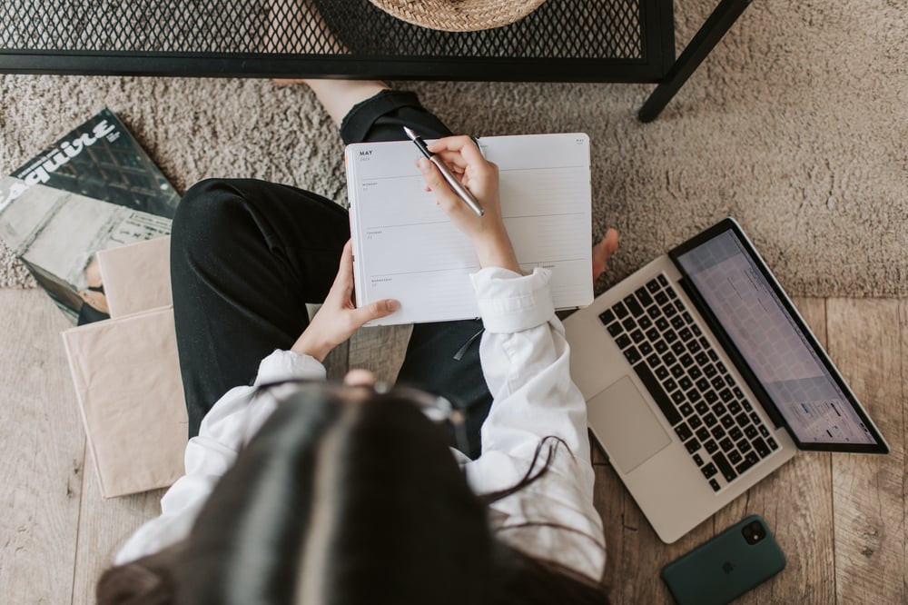 Overhead view of a writer sitting on the floor beside a laptop, writing in a planner.