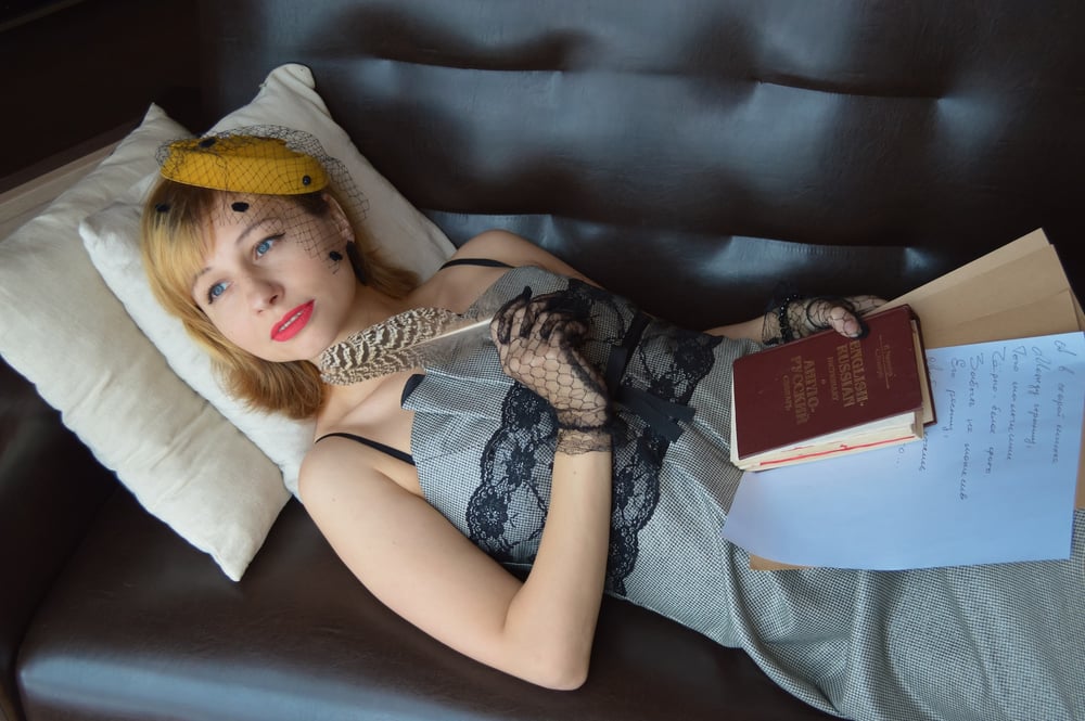 A female-presenting person in a dress and vintage yellow hat lies  a couch holding a quill, leather book, and stack of papers.