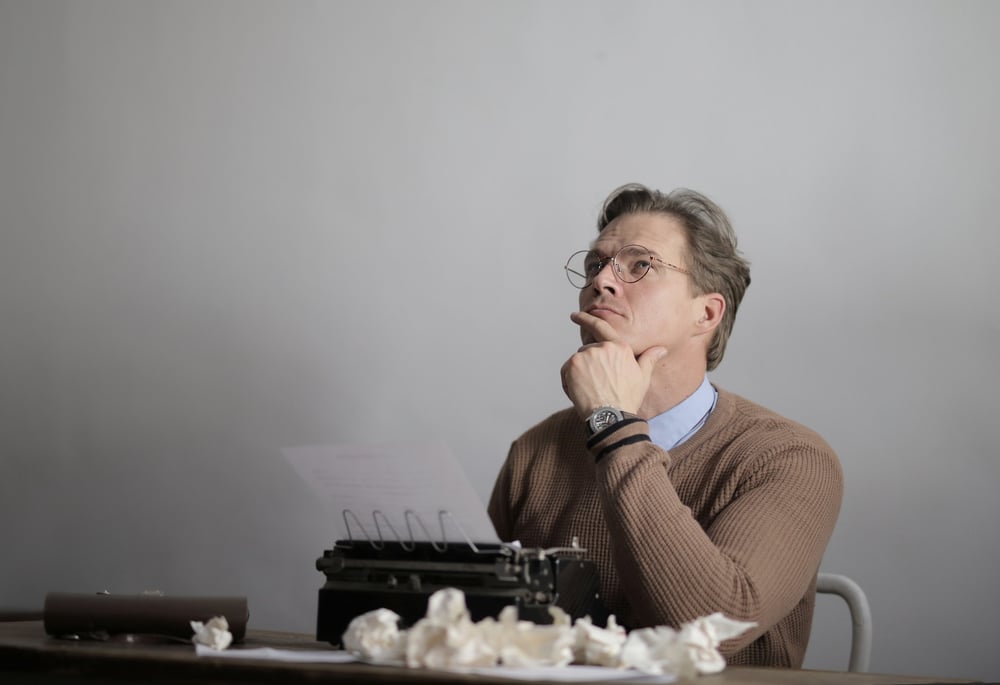 A writer sits thinking at a desk in front of a typewriter, wadded up paper strewn out in front of them.
