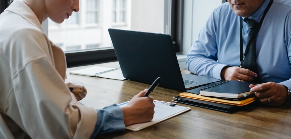 A person signs a contract at a desk while another person looks on.