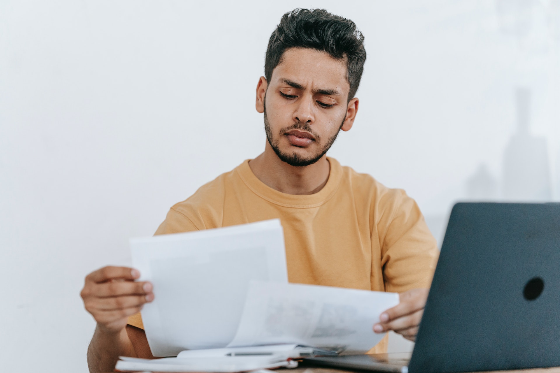 A male-presenting person sits in front of a laptop and examines a couple pieces of paper..