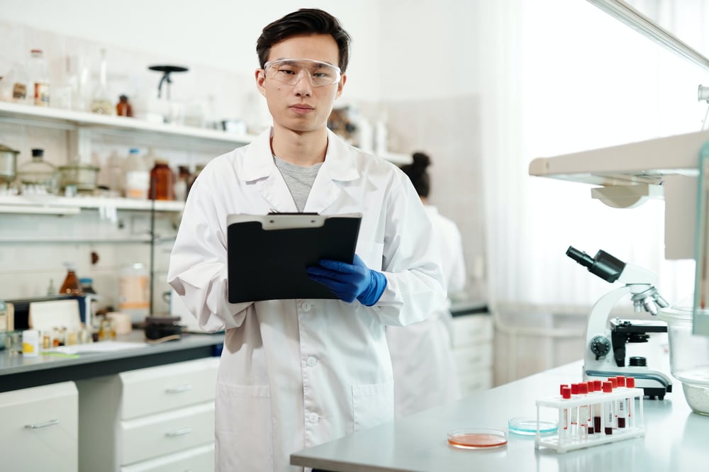 A scientist in a lab holds a clipboard and looks seriously at the camera through safety glasses.