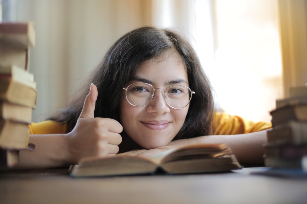 A person with glasses and long hair rests their chin on a book while giving a thumbs up.