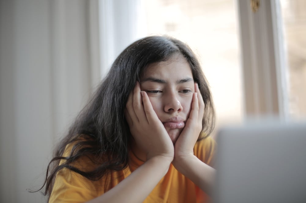 A person with long, dark hair rests their chin in their hands and looks at a computer screen with a disappointed expression.