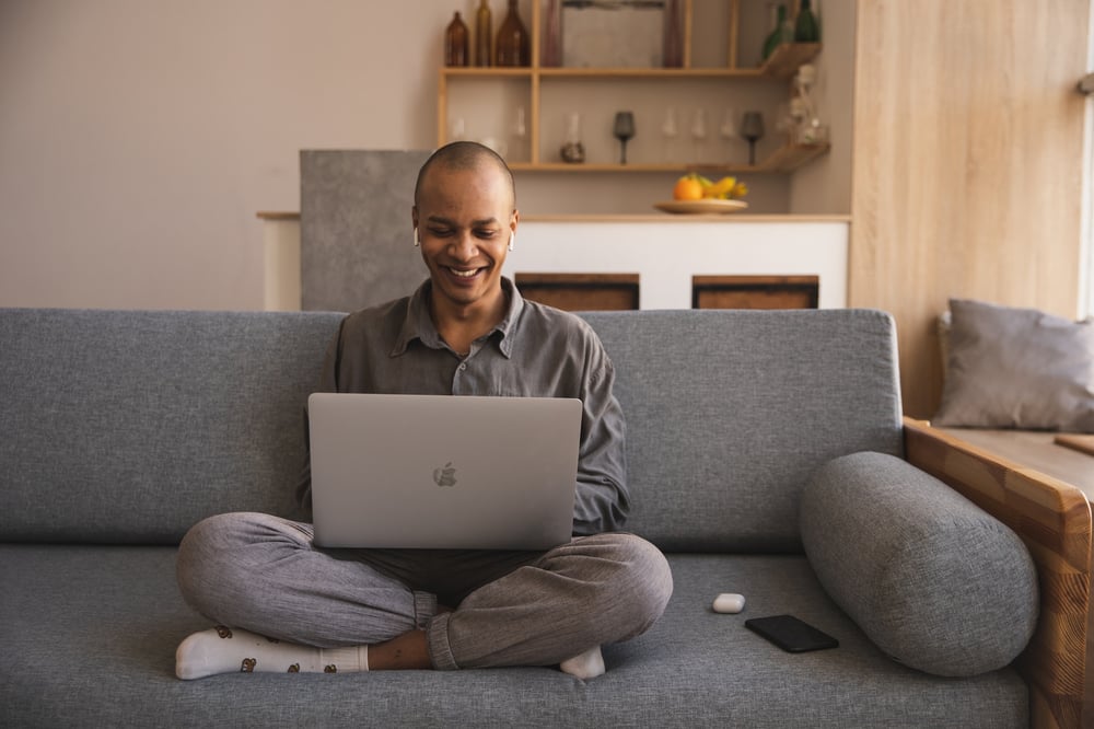 A smiling writer sits on a couch, typing on a laptop.