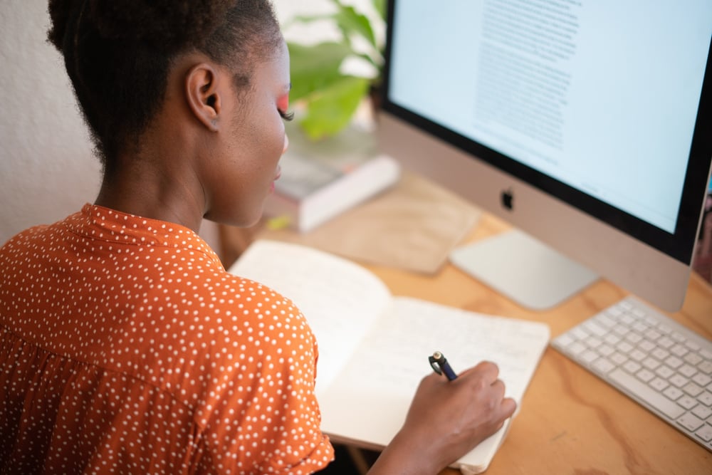 A writer sits in front of a big computer screen, writing in a notebook.