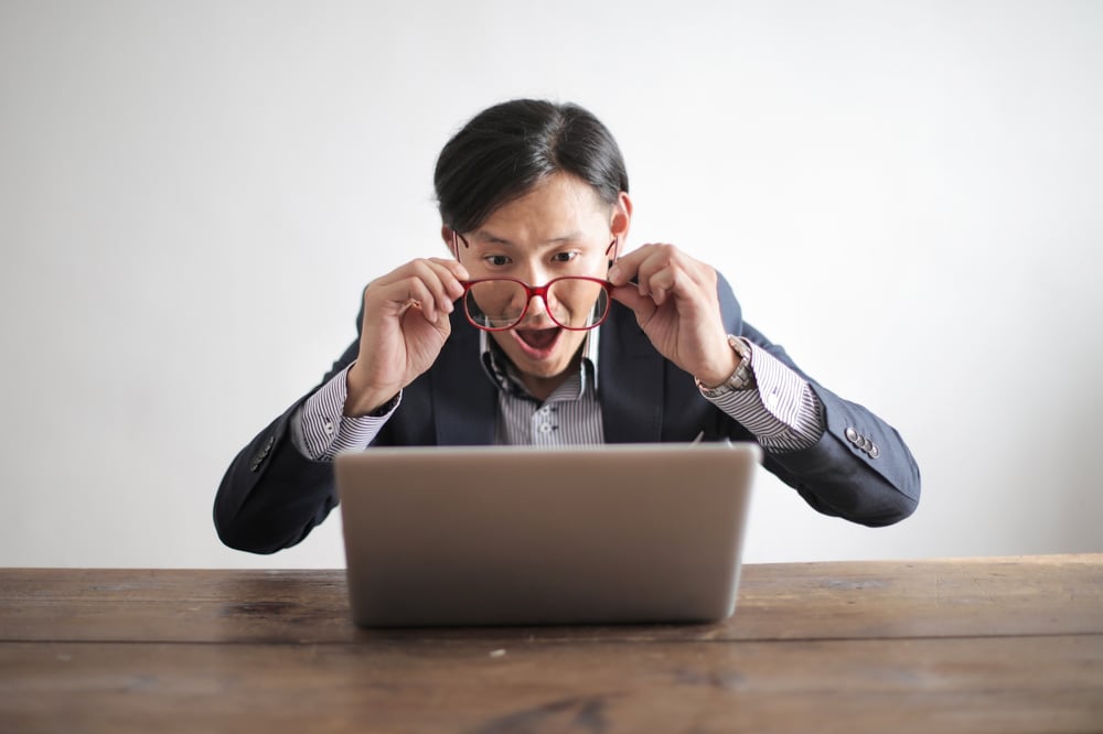 A writer in a suit lowers his glasses to look excitedly at a laptop screen.