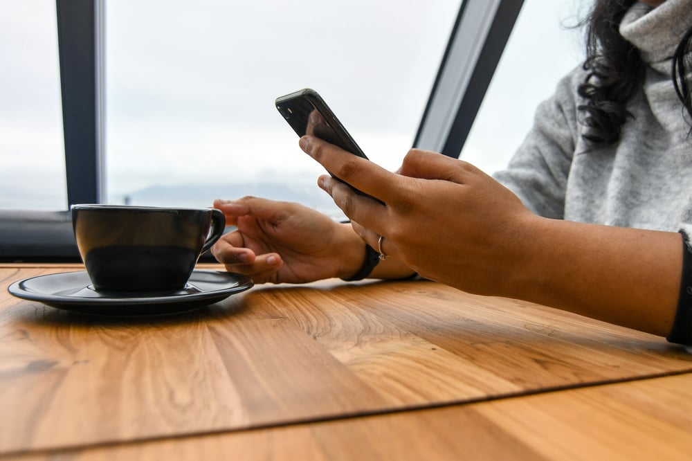 A person holds a phone in one hand and reaches for a cup of coffee with the other.