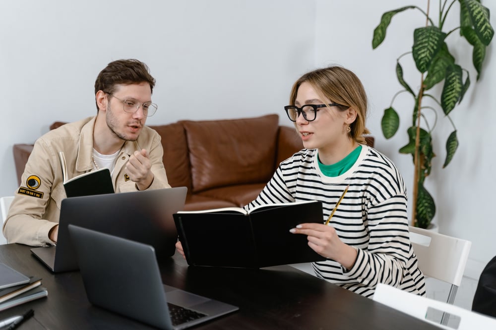 Two writers sit in front of open computers and look at notebooks, discussing something.