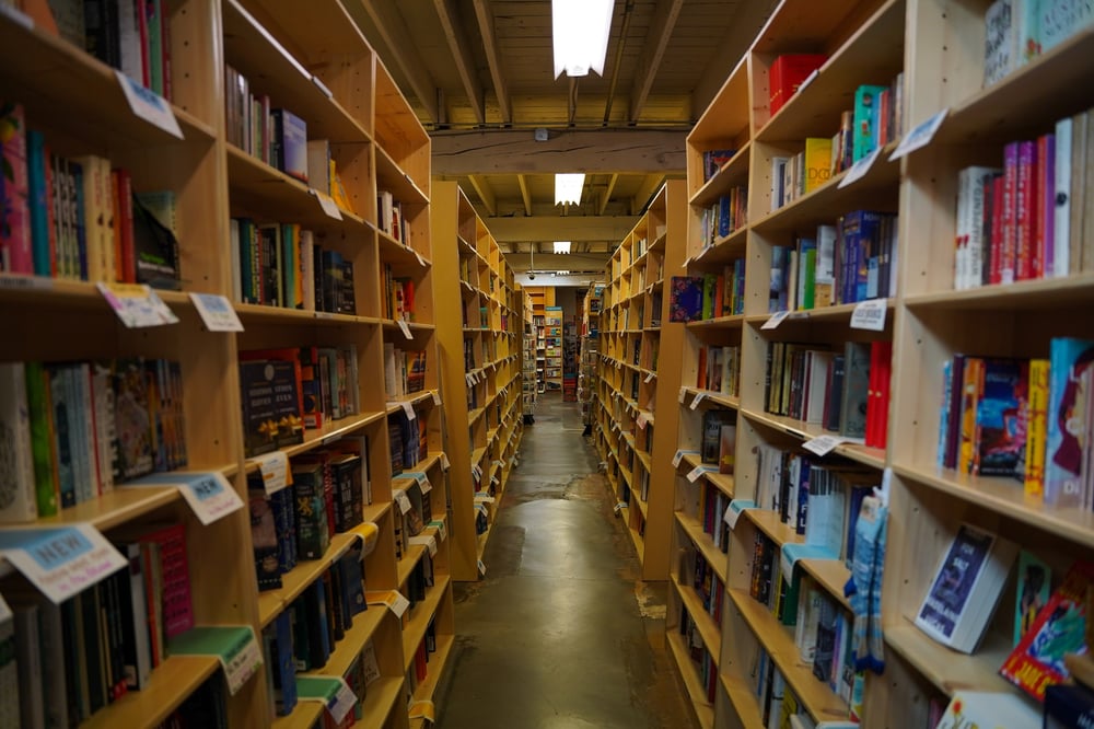 An aisle between two bookshelves that have literary genre labels on them.