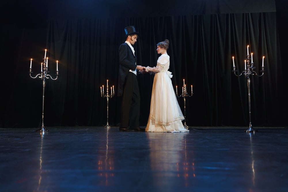 Two actors on a stage in historical costume and surrounded by giant candelabras.l