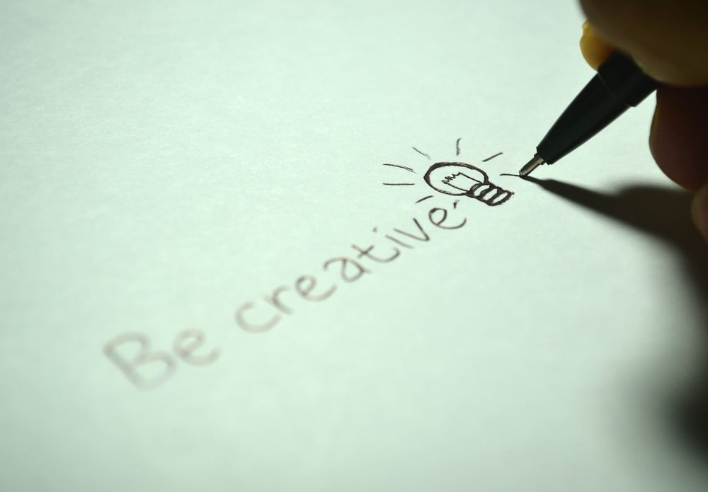 A hand writes "Be creative" and draws a light bulb on a white sheet of paper.