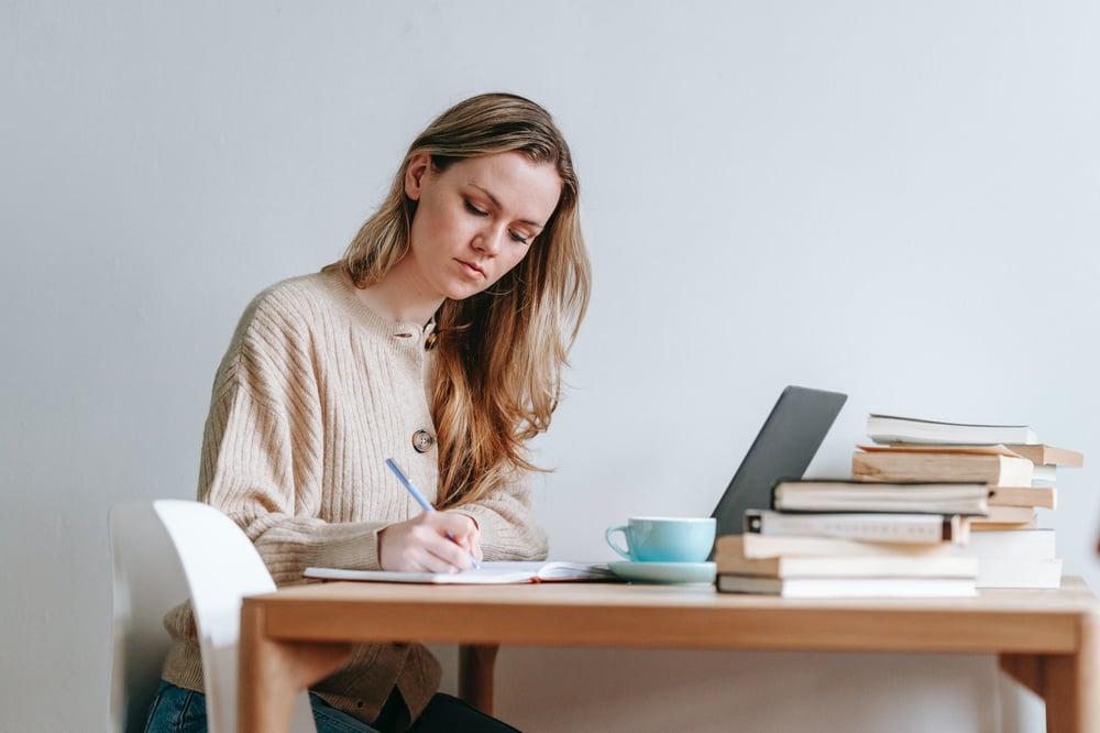 A person writes in a notebook beside a laptop and a stack of books.