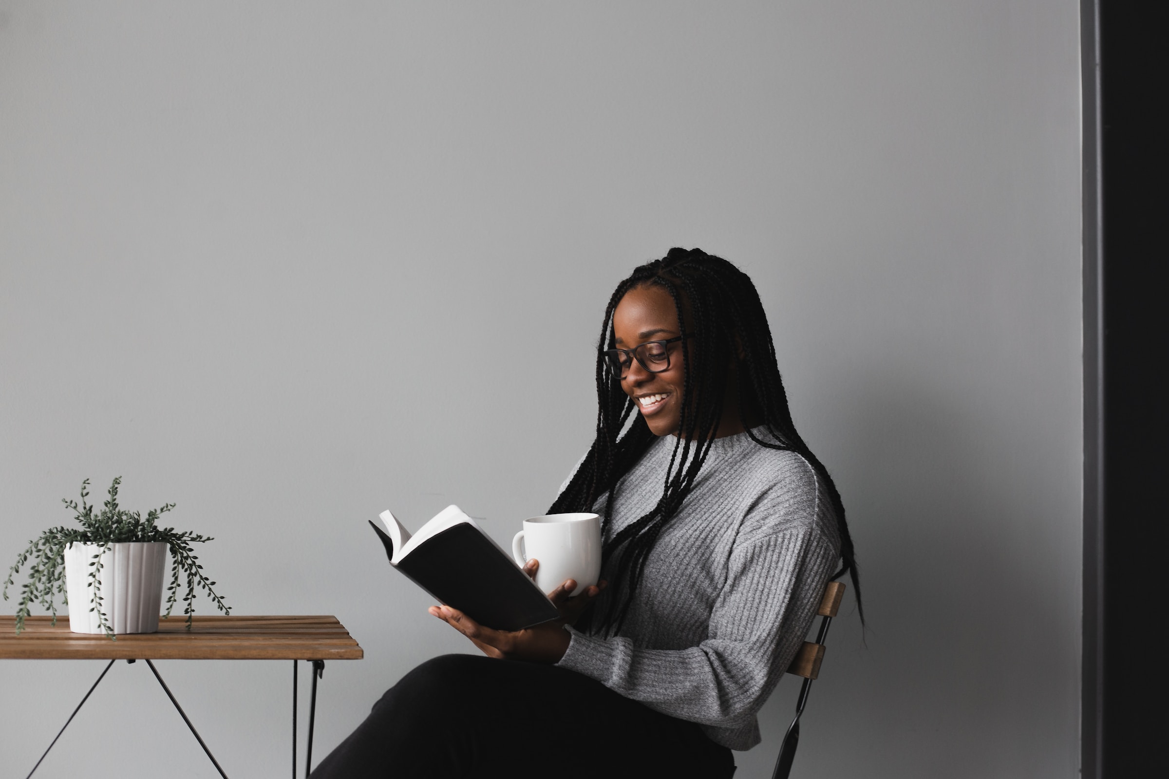 An adult reads a book while sitting at a small table, holding a cup of coffee.