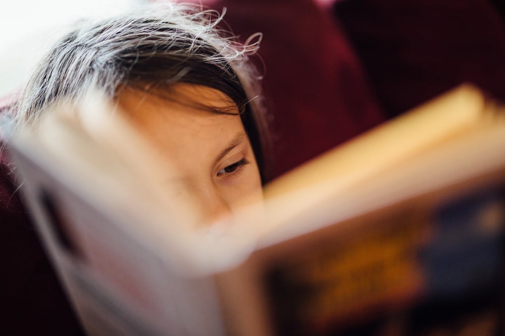 A young person's face barely visible over the top of the book they're reading.