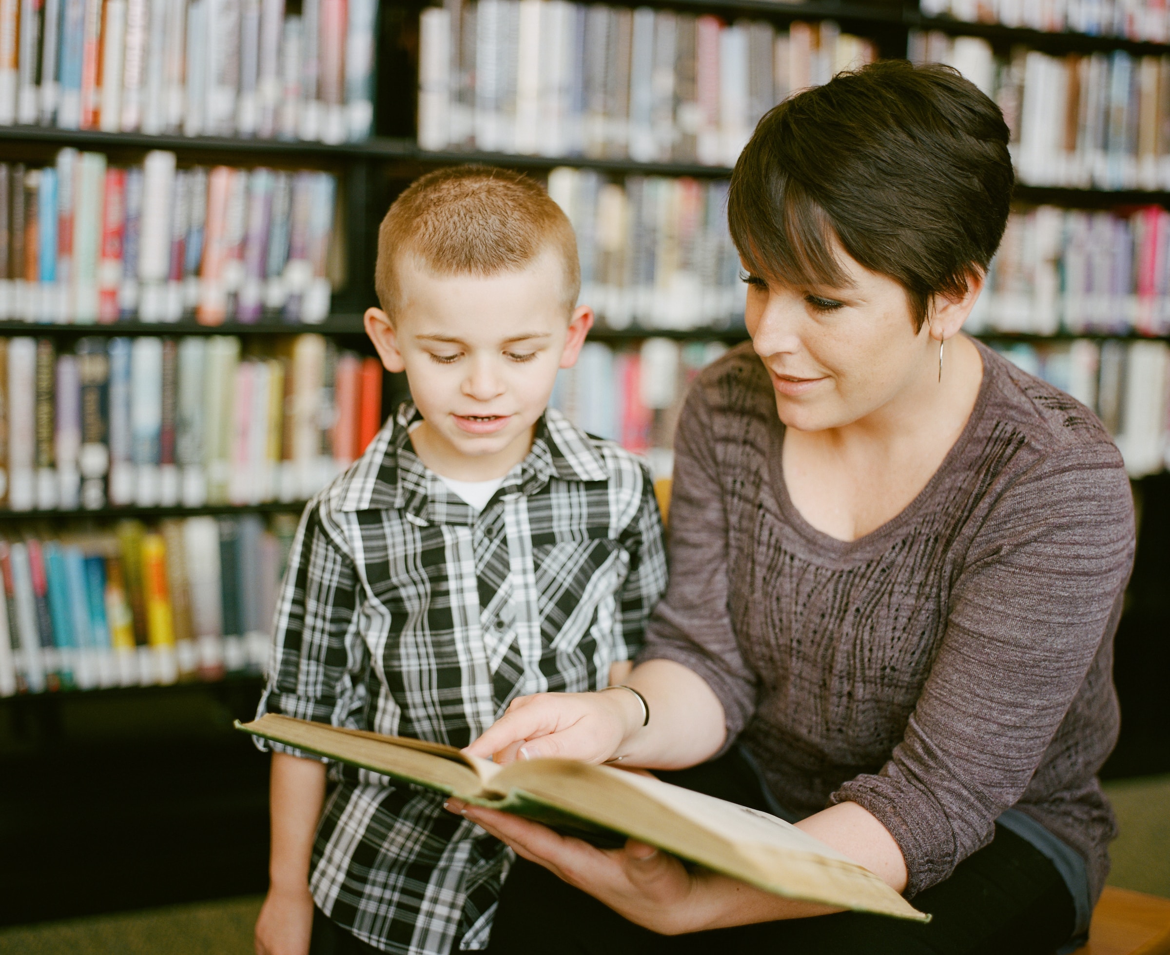A teacher helps a child read an early reader book in a library.