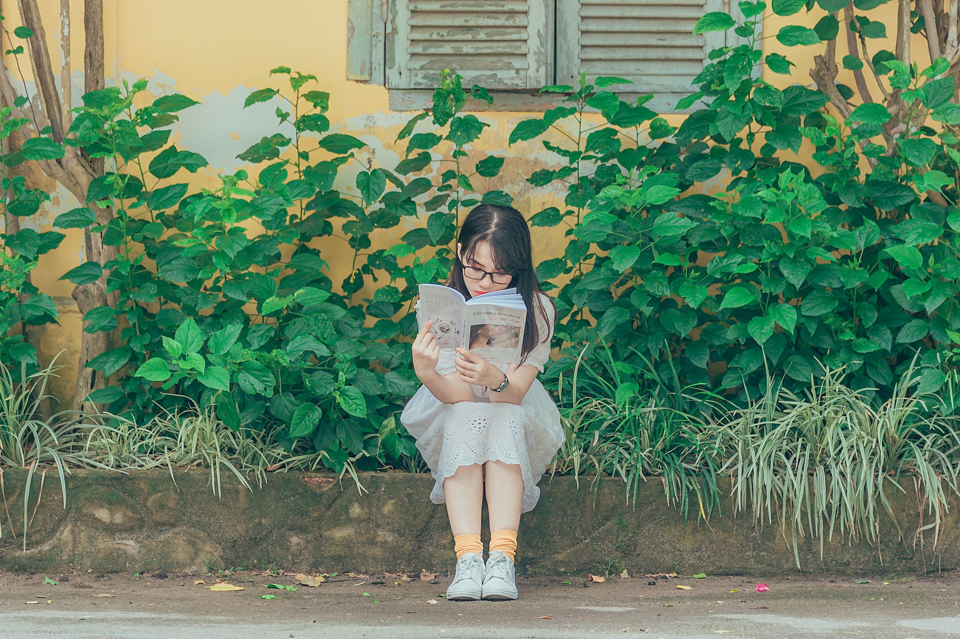A teenager in a white dress sits on a curb, reading a book.